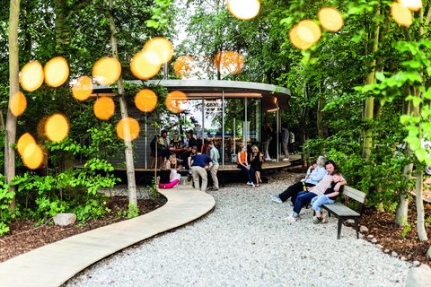 Belebter Garten um einen runden Pavillon; Menschen sitzen auf Bänken und am Steg, warme Lichtpunkte zwischen den Bäumen.