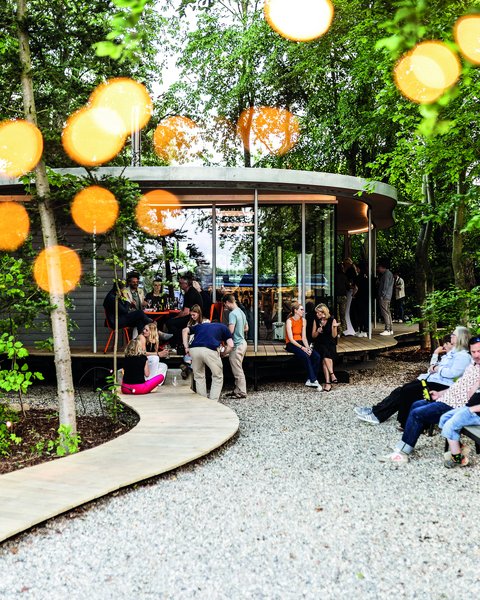 Belebter Garten um einen runden Pavillon; Menschen sitzen auf Bänken und am Steg, warme Lichtpunkte zwischen den Bäumen.