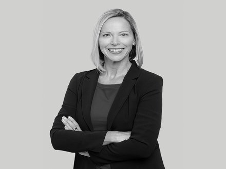 Schwarzweiß-Porträt, Frau vorn im Bild mit verschränkten Armen, freundlich lächelnd, dunkles Sakko und Oberteil, frontal aufgenommen vor einfarbig hellem Hintergrund.