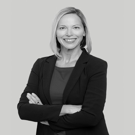 Schwarzweiß-Porträt, Frau vorn im Bild mit verschränkten Armen, freundlich lächelnd, dunkles Sakko und Oberteil, frontal aufgenommen vor einfarbig hellem Hintergrund.