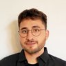 „Porträt eines jungen Mannes mit kurzen dunklen Haaren, Bart und runder Brille. Er trägt ein schwarzes Hemd und blickt freundlich in die Kamera.“