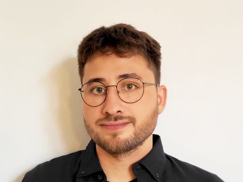 „Porträt eines jungen Mannes mit kurzen dunklen Haaren, Bart und runder Brille. Er trägt ein schwarzes Hemd und blickt freundlich in die Kamera.“
