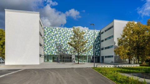 Moderner Schulgebäudekomplex mit weißer Fassade und mittigem Eingangsbereich mit grün-blau gemusterter Verkleidung, davor gepflasterter Platz und Bäume