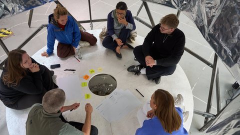 Sechs Personen sitzen im Kreis auf dem Boden um eine runde Öffnung mit Steinen und Papier mit Notizen und Stiften