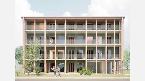Frontansicht eines viergeschossigen Holzbaus mit grüner Fassade: durchgehende Balkone mit Netzgeländern hinter Holzstützen; mehrere Personen auf den Laubengängen, Bäume davor.