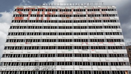 Breiter, leerstehender Plattenbau mit langen Reihen offener Fenster und Graffiti; oben der Schriftzug „ALLESANDERSPLATZ“ vor Wolkenhimmel.