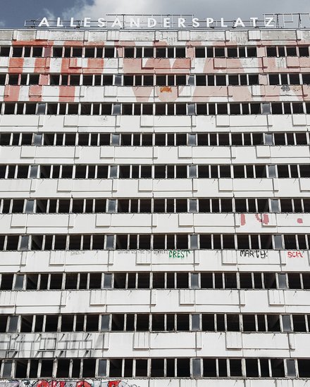 Breiter, leerstehender Plattenbau mit langen Reihen offener Fenster und Graffiti; oben der Schriftzug „ALLESANDERSPLATZ“ vor Wolkenhimmel.