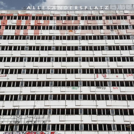 Breiter, leerstehender Plattenbau mit langen Reihen offener Fenster und Graffiti; oben der Schriftzug „ALLESANDERSPLATZ“ vor Wolkenhimmel.