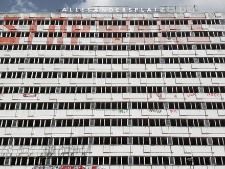 Breiter, leerstehender Plattenbau mit langen Reihen offener Fenster und Graffiti; oben der Schriftzug „ALLESANDERSPLATZ“ vor Wolkenhimmel.