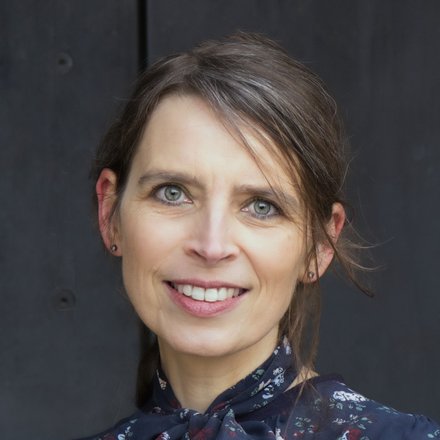 "Porträt einer lächelnden Frau mit zurückgebundenen Haaren. Sie trägt eine dunkelblaue Bluse mit Vogel- und Blumenmuster und steht vor einem dunkelgrauen Hintergrund."  Bild 3: Kochen im Freien