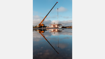 Kran hebt Bauteil auf einer Baustelle, gespiegelt in einer Wasserfläche.