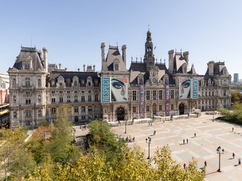 "Großer städtischer Platz vor einem imposanten Barockgebäude mit mehreren Türmen und aufwendiger Fassadengliederung. Das Rathaus oder Kulturgebäude trägt moderne Kunstbanner mit großformatigem Porträt. Der Platz ist teilweise gepflastert, mit einigen wenigen Menschen und grünen Bäumen in den Ecken. Modernes und historisches Erbe vereinen sich hier."