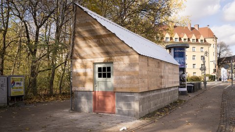 Kleines Holzhaus mit Satteldach auf massivem Sockel am Straßenrand; hellgrüne Tür, herbstliche Bäume, Litfaßsäule und Altbauten im Hintergrund.