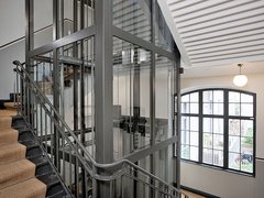 „Ein moderner Glasaufzug mit anthrazitfarbenem Stahlrahmen im Zentrum eines Treppenhauses mit Holzstufen und einem großen Sprossenfenster im Hintergrund.“