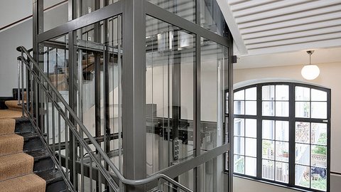 „Ein moderner Glasaufzug mit anthrazitfarbenem Stahlrahmen im Zentrum eines Treppenhauses mit Holzstufen und einem großen Sprossenfenster im Hintergrund.“