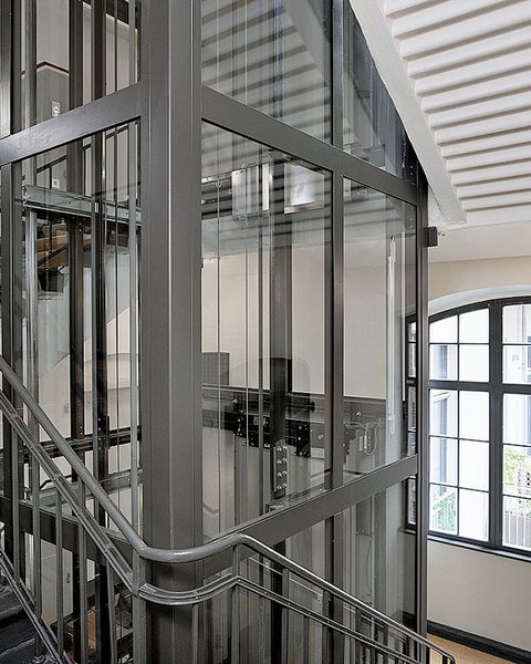 „Ein moderner Glasaufzug mit anthrazitfarbenem Stahlrahmen im Zentrum eines Treppenhauses mit Holzstufen und einem großen Sprossenfenster im Hintergrund.“