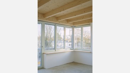 Helles Eckzimmer mit breiten Fenstern auf zwei Seiten und sichtbaren Holzdeckenbalken; durchgehende Fensterbank.