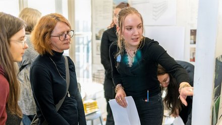 Mehrere Personen in einem hellen Büroraum. Eine Frau mit roten Haaren und Brille steht im Vordergrund und schaut nach links. Neben ihr eine junge Frau mit blonden Haaren in schwarzem Hemd, die ein Blatt Papier hält und lächelt. Im Hintergrund weitere Personen an großen Fenster mit Ausblick.
