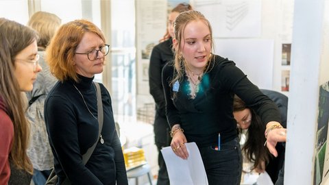 Mehrere Personen in einem hellen Büroraum. Eine Frau mit roten Haaren und Brille steht im Vordergrund und schaut nach links. Neben ihr eine junge Frau mit blonden Haaren in schwarzem Hemd, die ein Blatt Papier hält und lächelt. Im Hintergrund weitere Personen an großen Fenster mit Ausblick.