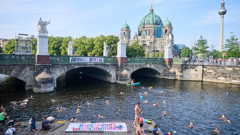 Zahlreiche Menschen schwimmen in einem Fluss unter einer Brücke in einer Großstadt. Im Hintergrund sind ein historischer Dom und ein Fernsehturm zu sehen, während Menschen das Geschehen vom Ufer aus beobachten.