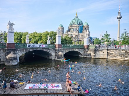 Zahlreiche Menschen schwimmen in einem Fluss unter einer Brücke in einer Großstadt. Im Hintergrund sind ein historischer Dom und ein Fernsehturm zu sehen, während Menschen das Geschehen vom Ufer aus beobachten.