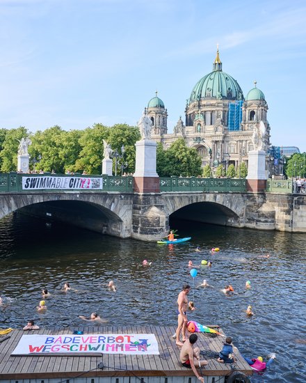 Zahlreiche Menschen schwimmen in einem Fluss unter einer Brücke in einer Großstadt. Im Hintergrund sind ein historischer Dom und ein Fernsehturm zu sehen, während Menschen das Geschehen vom Ufer aus beobachten.