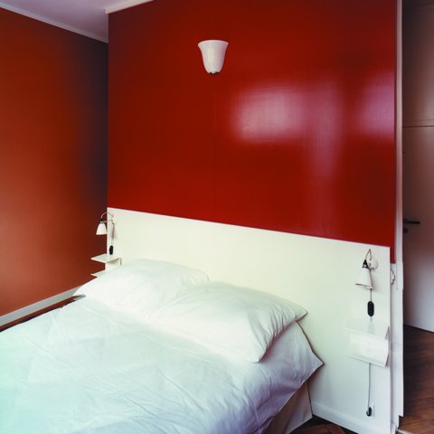 Schlafzimmer mit roter Wand, weißem Bett und kleiner Wandleuchte.