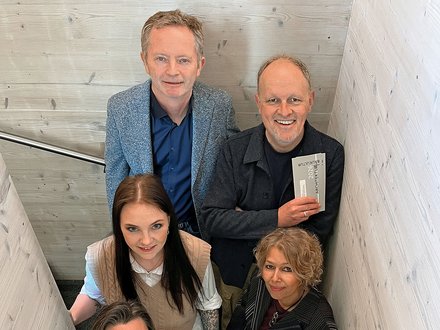 "Gruppenporträt von sechs Personen, angeordnet auf einer weißen Holztreppe. Oben links ein Mann mit grauem Haar in grauem Cardigan und blauem Hemd. Neben ihm ein Mann in schwarzer Jacke mit einem Dokument. Unten drei Frauen: eine mit schwarzem Haar und Brille in weißem Oberteil, eine mit blauem Hemd und beigefarbenem Accessoire, eine mit lockigem blondem Haar in schwarzer Pinstripe-Jacke. Alle blicken freundlich zur Kamera."