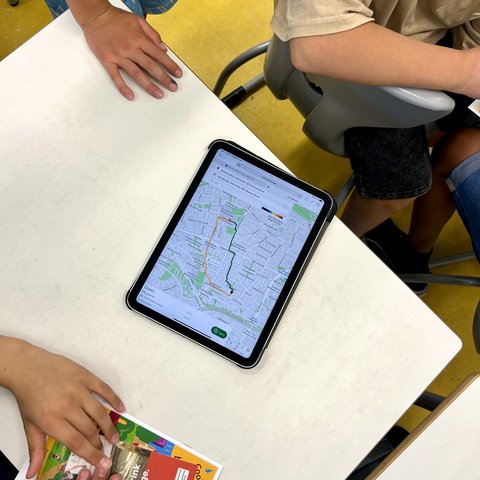 Blick von oben auf einen Tisch, auf dem ein iPad eine Karten-App mit zwei Routenoptionen anzeigt, umgeben von Händen mehrerer Personen.