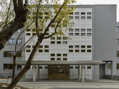 Fassade eines modernen Schulgebäudes mit rechteckigen Fenstern und einem überdachten Eingangsbereich, davor ein Baum mit grünen Blättern