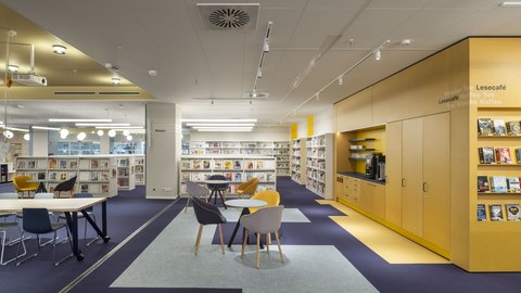 "Geräumige Erwachsenenbibliothek. Dunkelblaue Bodenzone mit hellen Teppichstreifen als Wegeführung. Links Bücherregale und Lesetische mit schwarzen und gelben Sesseln. Zentral mehrere Sessel (gelb, schwarz, grau) um eine kleine Teppichzone. Rechts gelbes Wandregal mit Bücherausstellung und "Lesecafé"-Bereich. Oben gelb gestrichene Nische mit Glasfront. Mehrere Bücherregale im Hintergrund. Weiße Decke mit linearer Beleuchtung. Großzügige, helle Atmosphäre."