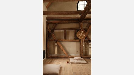 "Raum mit sichtbarem Holzfachwerk. Obere Wand aus heller Putzmasse. Rundes Fenster oben bringt Licht herein. Alte Holzstruktur mit diagonalen Kreuzbändern und Stützpfosten. Holzboden. Weiße Meditationskissen und -matten am Boden. Ein goldenes Becken hängt an der rechten Wand. Seildekorationen mit weißen Quasten."