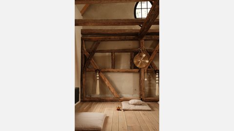 "Raum mit sichtbarem Holzfachwerk. Obere Wand aus heller Putzmasse. Rundes Fenster oben bringt Licht herein. Alte Holzstruktur mit diagonalen Kreuzbändern und Stützpfosten. Holzboden. Weiße Meditationskissen und -matten am Boden. Ein goldenes Becken hängt an der rechten Wand. Seildekorationen mit weißen Quasten."