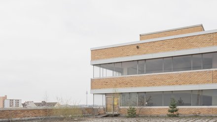 Dachfläche mit Blick über die Stadt  Eine flache Dachfläche mit hellem Belag und vereinzeltem Bewuchs. Im Vordergrund steht die Fassade eines Gebäudeteils mit Backstein und langen Fensterbändern. Im Hintergrund ist eine Stadtlandschaft mit einzelnen Hochpunkten zu erkennen. Das Bild zeigt das Gebäude als Teil der Stadt. Die Dachfläche wirkt ungenutzt, bietet aber Potenzial für neue Funktionen wie Aufenthalt, Begrünung oder Technik.