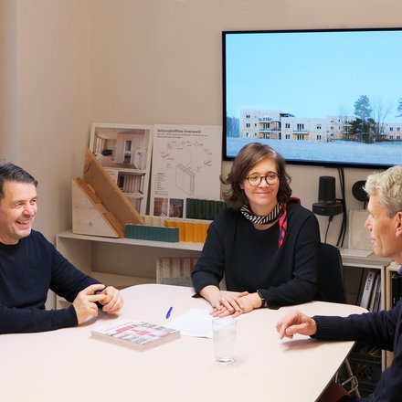 Drei Personen sitzen in einem Architekturbüro an einem Tisch vor Modellen und Bauplänen.