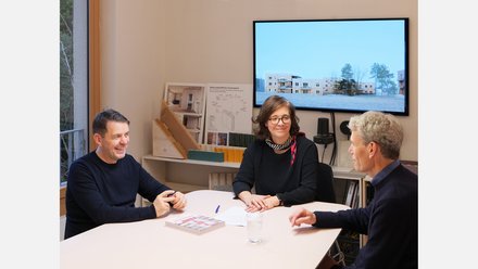 Drei Personen sitzen in einem Architekturbüro an einem Tisch vor Modellen und Bauplänen.