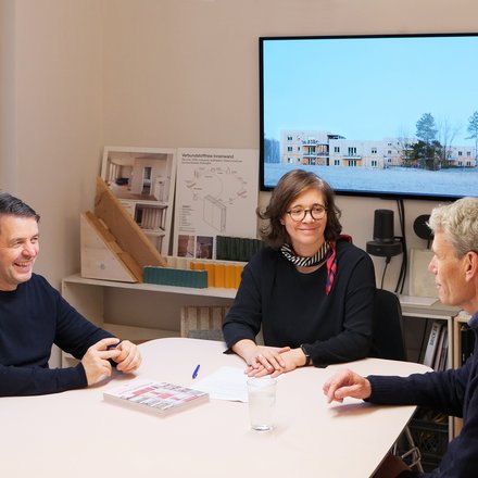 Drei Personen sitzen in einem Architekturbüro an einem Tisch vor Modellen und Bauplänen.