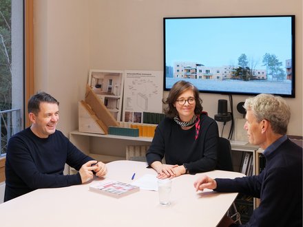 Drei Personen sitzen in einem Architekturbüro an einem Tisch vor Modellen und Bauplänen.
