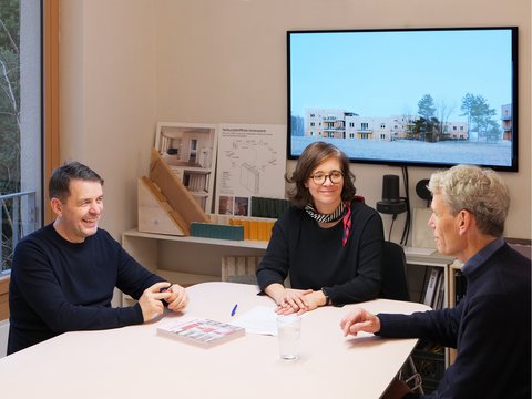 Drei Personen sitzen in einem Architekturbüro an einem Tisch vor Modellen und Bauplänen.
