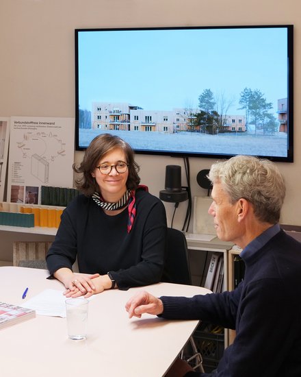 Drei Personen sitzen in einem Architekturbüro an einem Tisch vor Modellen und Bauplänen.