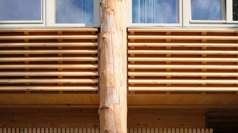Detail der Holzfassade: horizontale Lamellen, mittig eine runde Baumstammstütze unter einem Fensterband.