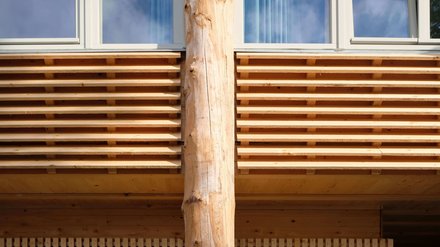 Detail der Holzfassade: horizontale Lamellen, mittig eine runde Baumstammstütze unter einem Fensterband.