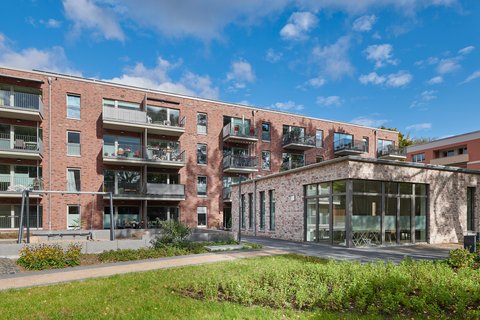  Ein moderner, vierstöckiger Wohnbau aus rötlichem Backstein unter blauem Himmel. Im Vordergrund erstreckt sich eine gepflegte Grünfläche mit einem kleinen Spielplatz (Schaukeln). Rechts ist ein flacher Anbau mit großen Glasfronten zu sehen, der Einblick in einen hellen Gemeinschaftsraum bietet. Die Hauptfassade verfügt über zahlreiche Balkone mit Metallgeländern.