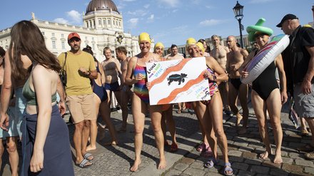 Eine Gruppe von Menschen in Badekleidung versammelt sich auf einem Platz vor einem historischen Gebäude. Zwei Personen halten ein Plakat für saubere und schwimmbare Flüsse in die Kamera.