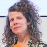  Frau mit schulterlangen, lockigen graubraunen Haaren, die ein schwarzes Oberteil mit buntem Blumenmuster trägt. Sie hält ein Mikrofon mit zwei farbigen Kugeln – eine orange und eine rosa – in den Händen. Sie blickt zur Seite und hat einen nachdenklichen Gesichtsausdruck. Der Hintergrund ist hellgrau.