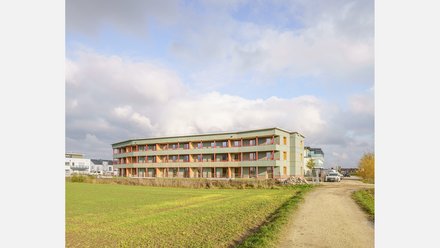 Langes, dreigeschossiges Wohnhaus mit grüner Holzverschalung und roten Akzenten am Feldrand unter bewölktem Himmel. Lang Gesamtansicht eines geschwungenen Mehrfamilienhauses mit grüner Holzfassade, durchlaufenden Balkonen/Laubengängen und roten Rahmen. Links Felder, rechts ein Feldweg mit geparkten Autos; Neubauten im Hintergrund.