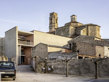 Historisches Steingebäude mit Kirchturm neben modernem, kubischem Neubau in ruhiger Hofsituation.