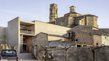 Historisches Steingebäude mit Kirchturm neben modernem, kubischem Neubau in ruhiger Hofsituation.