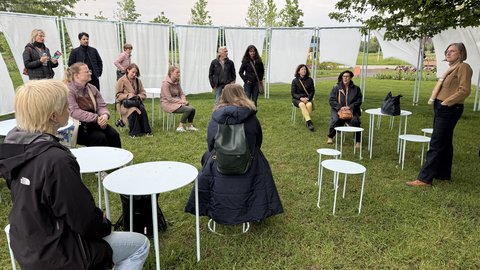  Eine Gruppe von etwa 15 Personen sitzt und steht auf einer grünen Wiese. Sie sind in einem Kreis angeordnet auf weißen Stühlen und verschiedenfarbigen Jacken. Im Hintergrund ist ein großes weißes Zelt mit Metallgestell gespannt. Der Himmel ist bewölkt. Die Szenerie deutet auf eine informelle Versammlung oder Veranstaltung hin.