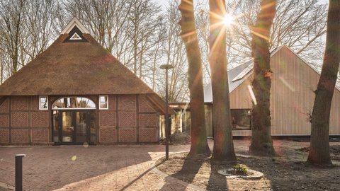 Das Foto zeigt ein Ensemble aus zwei Gebäuden: links ein traditionelles Fachwerkhaus mit Reetdach und bogenförmiger Glastür, rechts ein modernes Haus mit Holzfassade. Beide sind durch einen transparenten Gang verbunden und liegen in einem von Bäumen umgebenen Gelände. Ein gepflasterter Weg führt auf sie zu, die Sonne wirft lange Schatten.